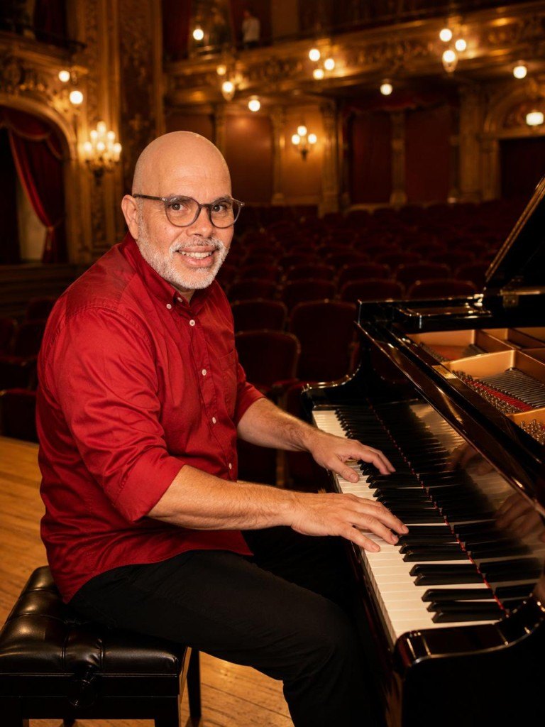 Alcir Alves ao piano, em um teatro, sorrindo para a câmera